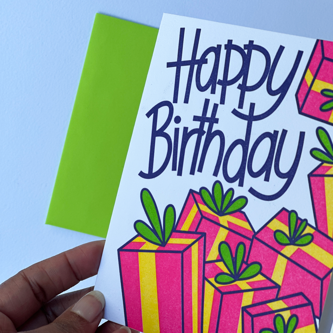 A hand with only a few fingers showing holding a Greeting Card with the words Happy Birthday and a design with a lot of gifts. The card is in a flat lay with a matching envelope.