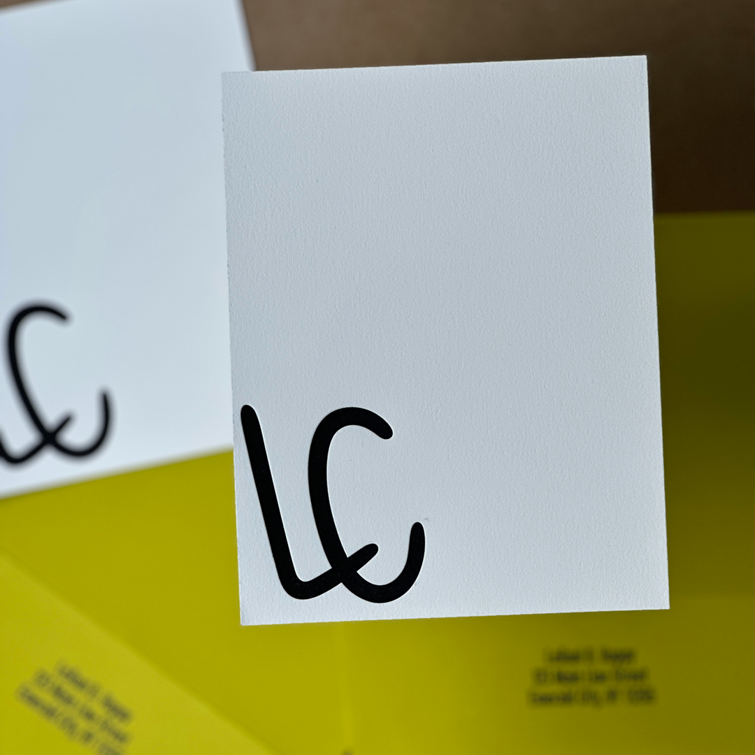 Close-up of a personalized letterpress stationery set featuring white cards with large, bold black initials 'LC' printed in the lower right corner. The cards are paired with yellow envelopes lined with black.