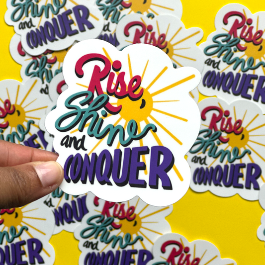 A hand holding an inspirational sticker with the words Rise, Shine and Conquer.