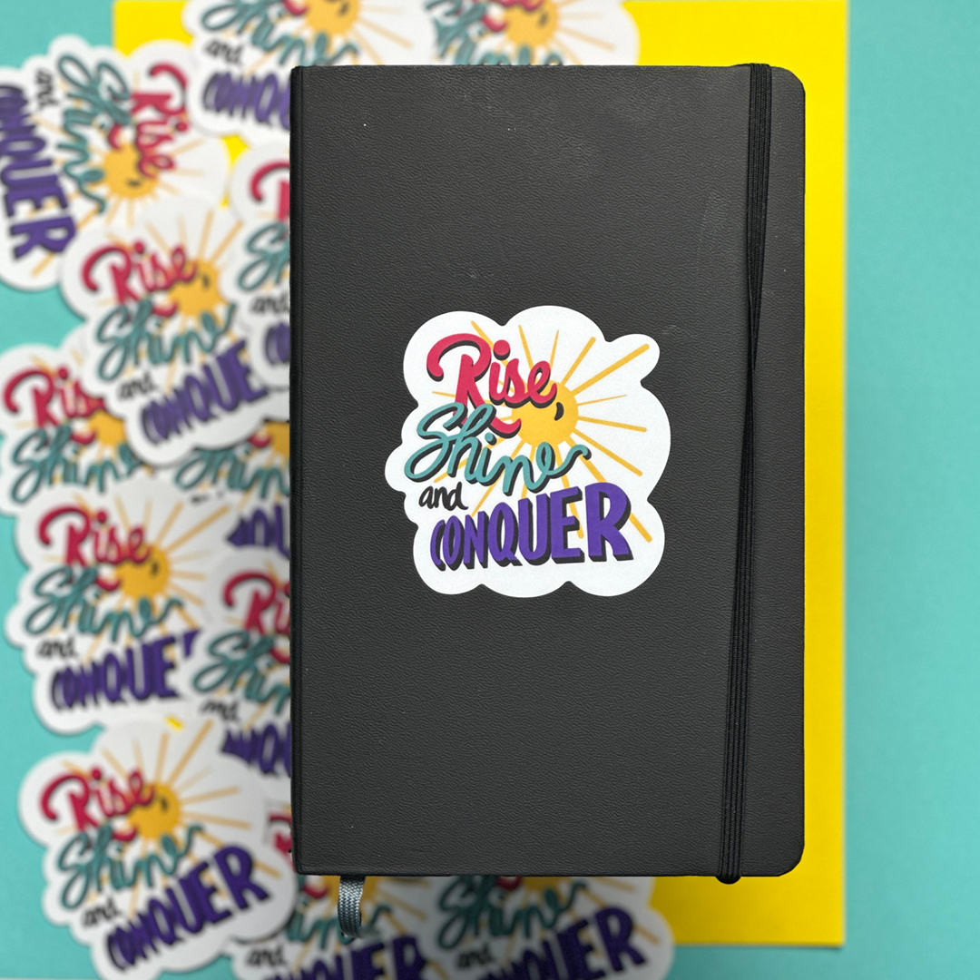 A sticker with the words Rise, Shine and Conquer on the front of a plain black journal.